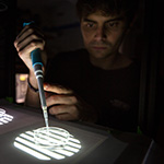 Marc Duque Ramirez (pictured), graduate student in the Harvard Neuroscience program, uses a large pipette to add liquid to a sample containing a days-old zebrafish. Chen and Duque Ramirez frequently work in their lab in near-total-darkness, to ensure that ambient light does not interfere with their findings.