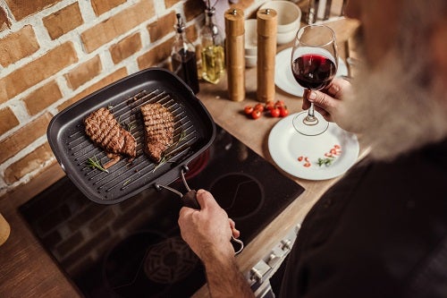 a man cooking steak on a griddle while holding a glass of wine
