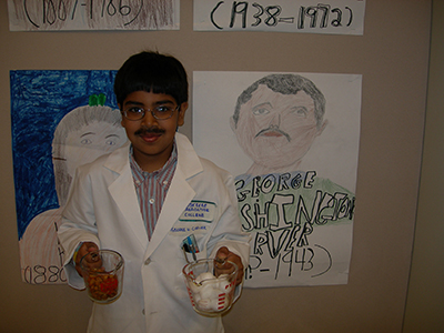 Akshay in the third grade dressed as George Washington Carver holding measuring cups with peanuts and cotton, two crops that Carver rotated to preserve soil health.