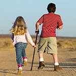 two children, a girl and a boy using a cane skip down a dirt road
