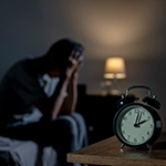 image depicting a tired person sitting up in bed with an alarm clock in the foreground